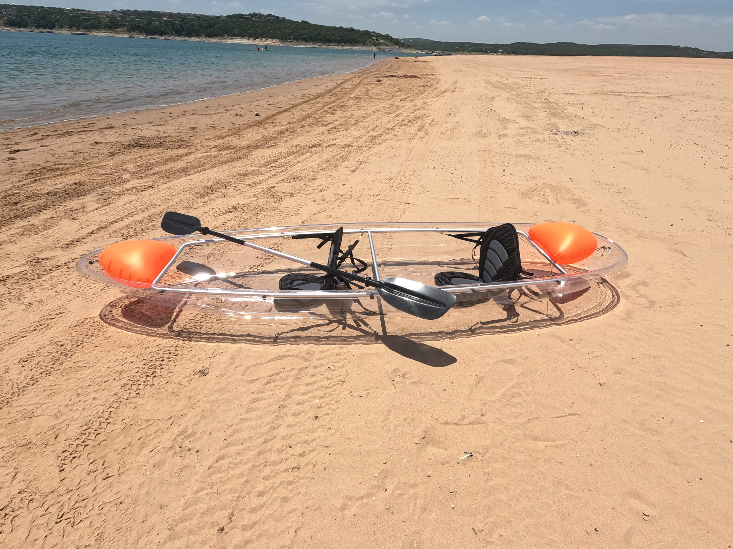 Full side profile of a clear tandem kayak with two black seats, a black paddle and orange flotation bags beside a lake.